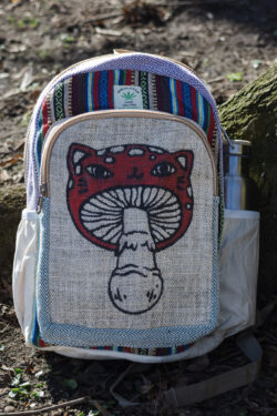 Kolorowy plecak leży na ziemi przy drzewie, ma beżowe kieszenie po bokach, jest wielkości A4 i wykonany z bawełny gheri oraz konopi, na przedniej kieszeni znajduje się ilustracja muchomora o podobiźnie kota