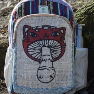 Kolorowy plecak leży na ziemi przy drzewie, ma beżowe kieszenie po bokach, jest wielkości A4 i wykonany z bawełny gheri oraz konopi, na przedniej kieszeni znajduje się ilustracja muchomora o podobiźnie kota