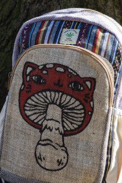 Kolorowy plecak leży na ziemi przy drzewie, ma beżowe kieszenie po bokach, jest wielkości A4 i wykonany z bawełny gheri oraz konopi, na przedniej kieszeni znajduje się ilustracja muchomora o podobiźnie kota