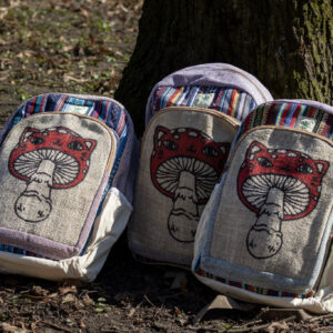 Trzy kolorowe plecaki leżą na ziemi przy drzewie, mają beżowe kieszenie po bokach, są wielkości A4 i wykonane z bawełny gheri oraz konopi, na przedniej kieszeni znajduje się ilustracja muchomora o podobiźnie kota