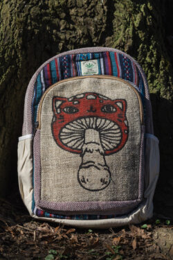 Kolorowy plecak leży na ziemi przy drzewie, ma beżowe kieszenie po bokach, jest wielkości A4 i wykonany z bawełny gheri oraz konopi, na przedniej kieszeni znajduje się ilustracja muchomora o podobiźnie kota