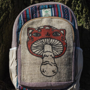 Kolorowy plecak leży na ziemi przy drzewie, ma beżowe kieszenie po bokach, jest wielkości A4 i wykonany z bawełny gheri oraz konopi, na przedniej kieszeni znajduje się ilustracja muchomora o podobiźnie kota