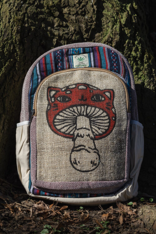 Kolorowy plecak leży na ziemi przy drzewie, ma beżowe kieszenie po bokach, jest wielkości A4 i wykonany z bawełny gheri oraz konopi, na przedniej kieszeni znajduje się ilustracja muchomora o podobiźnie kota