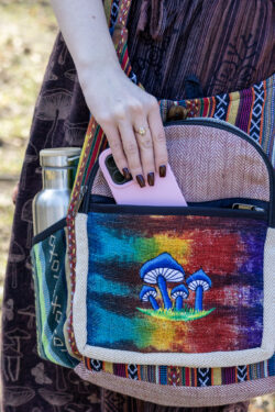 Osoba prezentuje kolorową torbę wykonaną z materiału gheri i konopi, wkłada telefon do przedniej kieszeni; w bocznej kieszeni znajduje się srebrny bidon, w tle trawa