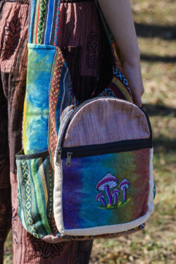 Kobieta w czerwonych patchworkowych spodniach i czarnym topie prezentuje tęczową torbę z ilustracją grzybów, noszoną na ramieniu, wykonaną z materiału gheri i konopi, na tle trawy