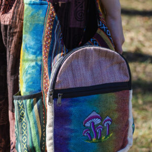 Kobieta w czerwonych patchworkowych spodniach i czarnym topie prezentuje tęczową torbę z ilustracją grzybów, noszoną na ramieniu, wykonaną z materiału gheri i konopi, na tle trawy