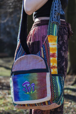Kobieta w czerwonych patchworkowych spodniach i czarnym topie prezentuje tęczową torbę z ilustracją grzybów, noszoną na ramieniu, wykonaną z materiału gheri i konopi, na tle trawy