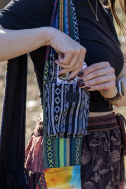 Osoba w czerwonych spodniach i czarnym topie wkłada biały telefon do małej kieszeni torby noszonej na ramieniu, we wzór gheri w kolorach czerni i bieli, zamykanej na czarny rzep, na tle trawy