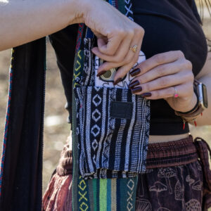 Osoba w czerwonych spodniach i czarnym topie wkłada biały telefon do małej kieszeni torby noszonej na ramieniu, we wzór gheri w kolorach czerni i bieli, zamykanej na czarny rzep, na tle trawy