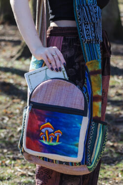 Kobieta w czerwonych patchworkowych spodniach i czarnym topie wkłada biały notes do przedniej kieszeni kolorowej torby z ilustracją grzybów w dominujących kolorach czerwieni i niebieskiego, noszonej na ramieniu, wykonanej z materiału gheri i konopi, na tle trawy