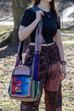 Kobieta w czerwonych patchworkowych spodniach i czarnym topie prezentuje kolorową torbę z ilustracją grzybów w dominujących kolorach czerwieni i niebieskiego, noszoną na ramieniu, wykonaną z materiału gheri i konopi, na tle trawy