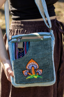 Osoba trzymająca zieloną, kolorową torebkę z wzorem pomarańczowych grzybów, ubrana w brązowe szarawary i czarny top, na tle trawy