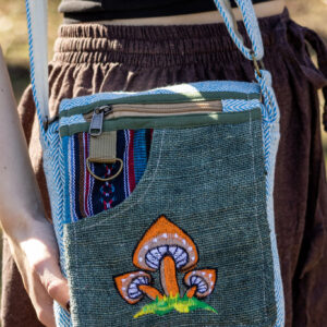 Osoba trzymająca zieloną, kolorową torebkę z wzorem pomarańczowych grzybów, ubrana w brązowe szarawary i czarny top, na tle trawy