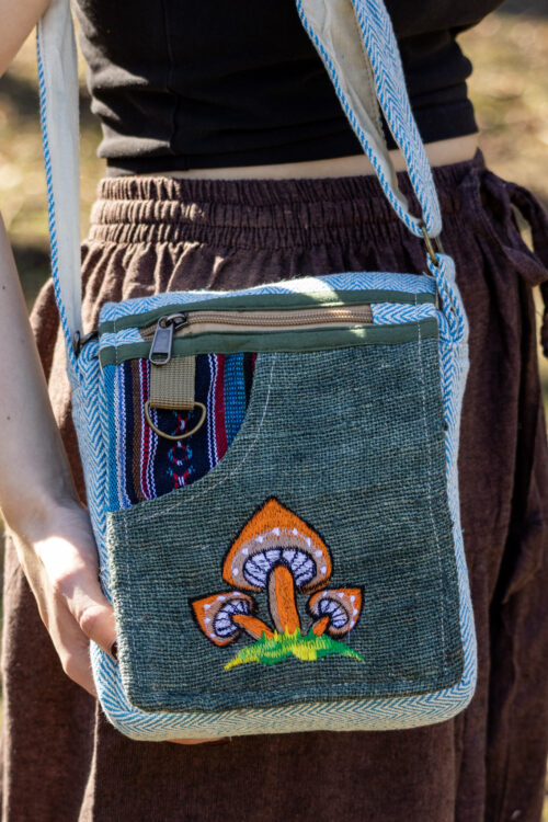 Osoba trzymająca zieloną, kolorową torebkę z wzorem pomarańczowych grzybów, ubrana w brązowe szarawary i czarny top, na tle trawy