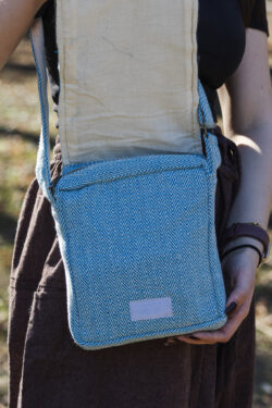 kobieta w brązowych szarawarach i czarnym topie otwiera klapę torebki w niebiesko-biały wzór jodełki z białym zapięciem na rzep, na tle trawy