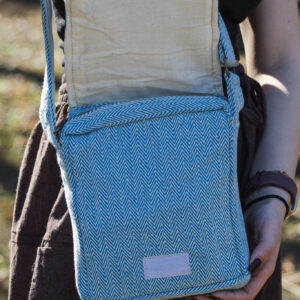 kobieta w brązowych szarawarach i czarnym topie otwiera klapę torebki w niebiesko-biały wzór jodełki z białym zapięciem na rzep, na tle trawy