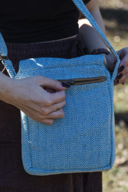 Osoba w brązowych szarawarach i czarnym topie zasuwa tylną kieszeń torebki w niebiesko-biały wzór jodełki, na tle trawy