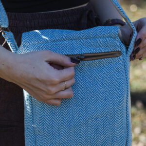 Osoba w brązowych szarawarach i czarnym topie zasuwa tylną kieszeń torebki w niebiesko-biały wzór jodełki, na tle trawy