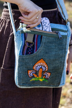 Osoba w brązowych szarawarach i czarnym topie wkłada telefon do zapinanej kieszeni zielonej torebki z motywem pomarańczowych grzybów, na tle trawy