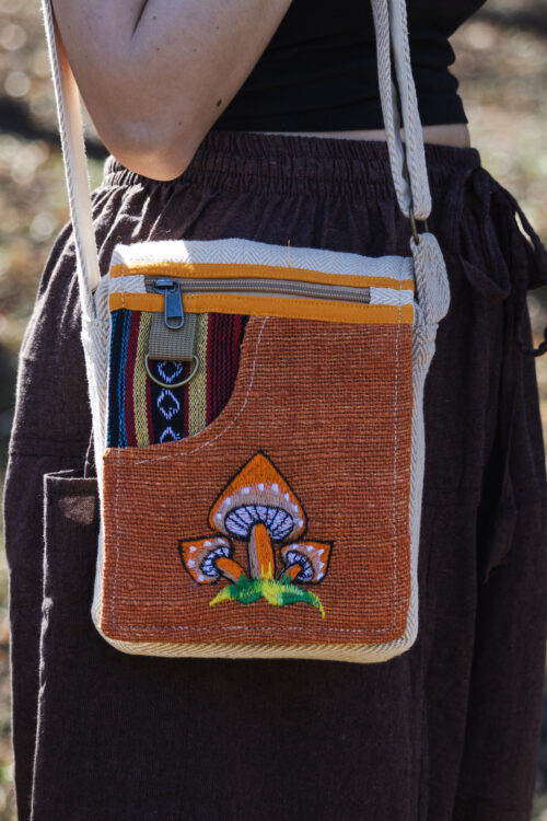 Osoba nosząca pomarańczową, kolorową torebkę z wzorem grzybów, ubrana w brązowe szarawary i czarny top, na tle trawy.