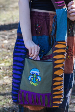 Osoba w spodniach w etniczne kolorowe wzory i czarnym topie wkłada telefon do przedniej kieszeni torby dominujący kolor kieszeni to zielony z ilustracją niebieskich grzybów torba patchwork z rozcięciami