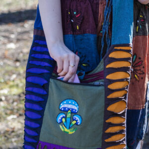 Osoba w spodniach w etniczne kolorowe wzory i czarnym topie wkłada telefon do przedniej kieszeni torby dominujący kolor kieszeni to zielony z ilustracją niebieskich grzybów torba patchwork z rozcięciami