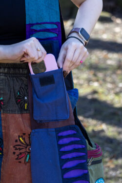 Osoba wkłada telefon do kieszonki na rzep na pasku torby ma na sobie patchworkowe spodnie w etniczne wzory i czarny top na tle trawy