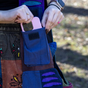 Osoba wkłada telefon do kieszonki na rzep na pasku torby ma na sobie patchworkowe spodnie w etniczne wzory i czarny top na tle trawy