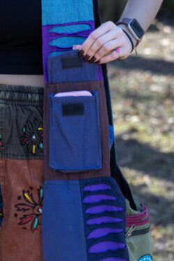 Osoba otwiera kieszonkę na rzep na pasku torby w której znajduje się telefon ma na sobie patchworkowe spodnie w etniczne wzory i czarny top na tle trawy