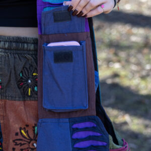 Osoba otwiera kieszonkę na rzep na pasku torby w której znajduje się telefon ma na sobie patchworkowe spodnie w etniczne wzory i czarny top na tle trawy