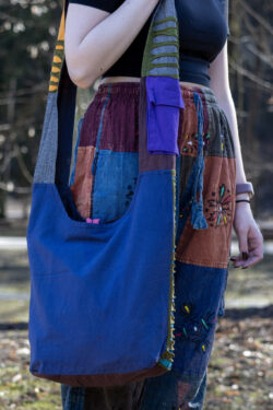 Osoba w kolorowych spodniach i czarnym topie prezentuje tył torby na ramieniu w niebieskim kolorze z fioletową kieszonką na pasku