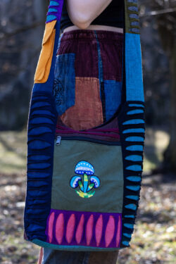 Osoba w kolorowych spodniach ma na ramieniu torbę patchwork z ozdobnymi rozcięciami i ilustracją niebieskich grzybów w parku