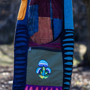 Osoba w kolorowych spodniach ma na ramieniu torbę patchwork z ozdobnymi rozcięciami i ilustracją niebieskich grzybów w parku