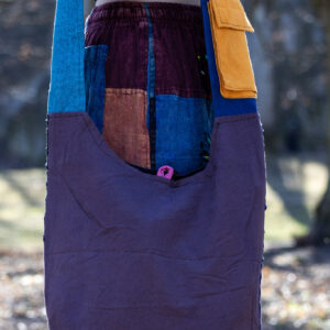 Osoba w kolorowych spodniach i czarnym topie prezentuje tył torby na ramieniu w niebieskim kolorze z fioletową kieszonką na pasku