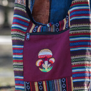 Osoba w kolorowych spodniach ma na ramieniu torbę z materiału gheri i ilustracją pomarańczowych grzybów na tle trawy