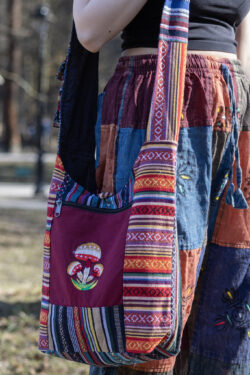 Osoba w kolorowych spodniach ma na ramieniu torbę z materiału gheri i ilustracją pomarańczowych grzybów na tle trawy