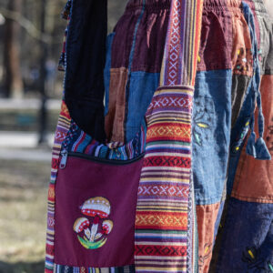 Osoba w kolorowych spodniach ma na ramieniu torbę z materiału gheri i ilustracją pomarańczowych grzybów na tle trawy