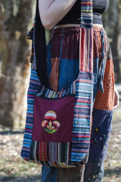 Osoba w kolorowych spodniach ma na ramieniu torbę z materiału gheri i ilustracją pomarańczowych grzybów na tle trawy