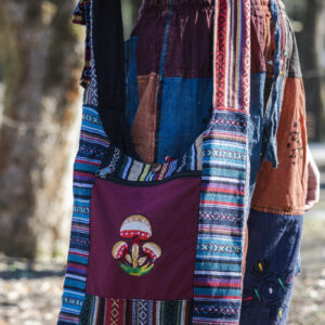 Osoba w kolorowych spodniach ma na ramieniu torbę z materiału gheri i ilustracją pomarańczowych grzybów na tle trawy