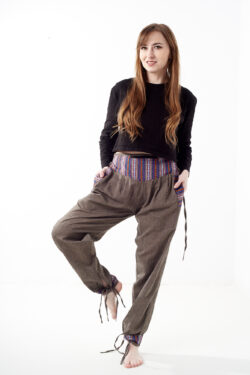 Kobieta na białym tle w czarnym topie i brązowych, luźnych bawełnianych spodniach z wiązaniami na nogawkach oraz wykończeniami we wzór gheri, z przodu znajduje się rozpinana kieszeń