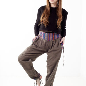 Kobieta na białym tle w czarnym topie i brązowych, luźnych bawełnianych spodniach z wiązaniami na nogawkach oraz wykończeniami we wzór gheri, z przodu znajduje się rozpinana kieszeń