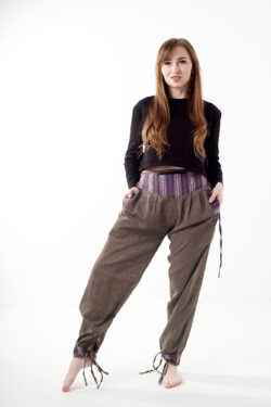 Kobieta na białym tle w czarnym topie i brązowych, luźnych bawełnianych spodniach z wiązaniami na nogawkach oraz wykończeniami we wzór gheri, z przodu znajduje się rozpinana kieszeń