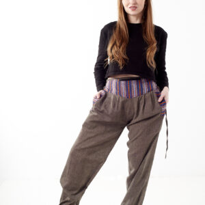 Kobieta na białym tle w czarnym topie i brązowych, luźnych bawełnianych spodniach z wiązaniami na nogawkach oraz wykończeniami we wzór gheri, z przodu znajduje się rozpinana kieszeń