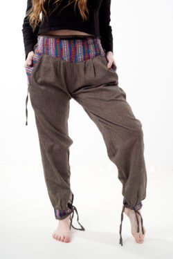 Kobieta na białym tle w czarnym topie i brązowych, luźnych bawełnianych spodniach z wiązaniami na nogawkach oraz wykończeniami we wzór gheri, z przodu znajduje się rozpinana kieszeń