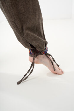 Zbliżenie na nogawkę brązowych, luźnych bawełnianych spodni z wiązaniem i wykończeniem we wzór gheri, na białym tle.