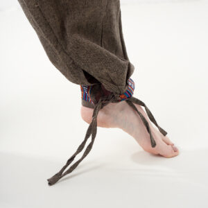 Zbliżenie na nogawkę brązowych, luźnych bawełnianych spodni z wiązaniem i wykończeniem we wzór gheri, na białym tle.