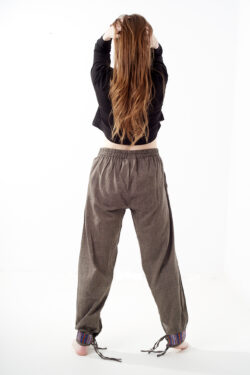 Kobieta na białym tle stoi tyłem w czarnym topie i brązowych, luźnych bawełnianych spodniach z wiązaniami na nogawkach oraz wykończeniami we wzór gheri