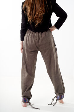 Kobieta na białym tle stoi tyłem w czarnym topie i brązowych, luźnych bawełnianych spodniach z wiązaniami na nogawkach oraz wykończeniami we wzór gheri