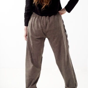 Kobieta na białym tle stoi tyłem w czarnym topie i brązowych, luźnych bawełnianych spodniach z wiązaniami na nogawkach oraz wykończeniami we wzór gheri