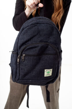 Osoba na białym tle w brązowych spodniach we wzór gheri i czarnym topie trzyma w rękach czarny plecak z konopi z wieloma zapięciami